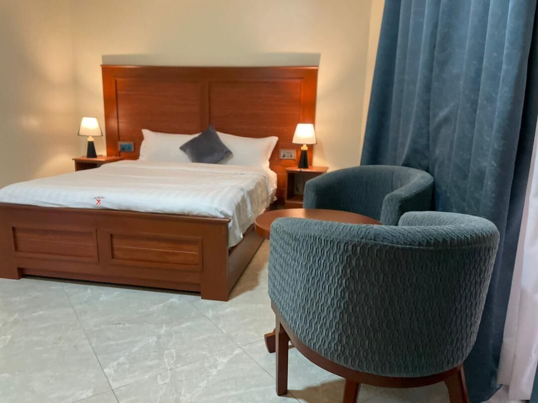 A hotel room with a wooden bed, two bedside tables with lamps, a blue armchair, and a large window with curtains.