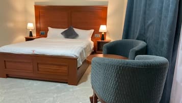 A hotel room with a wooden bed, two bedside tables with lamps, a blue armchair, and a large window with curtains.