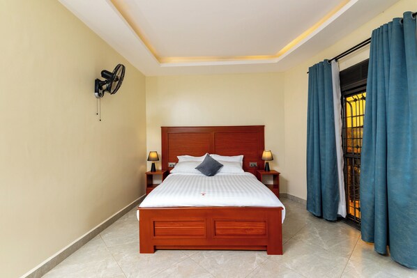 A neatly made bed with a wooden headboard, two bedside lamps, and a wall-mounted fan. - Kaije Country Cottages (Fort Portal)