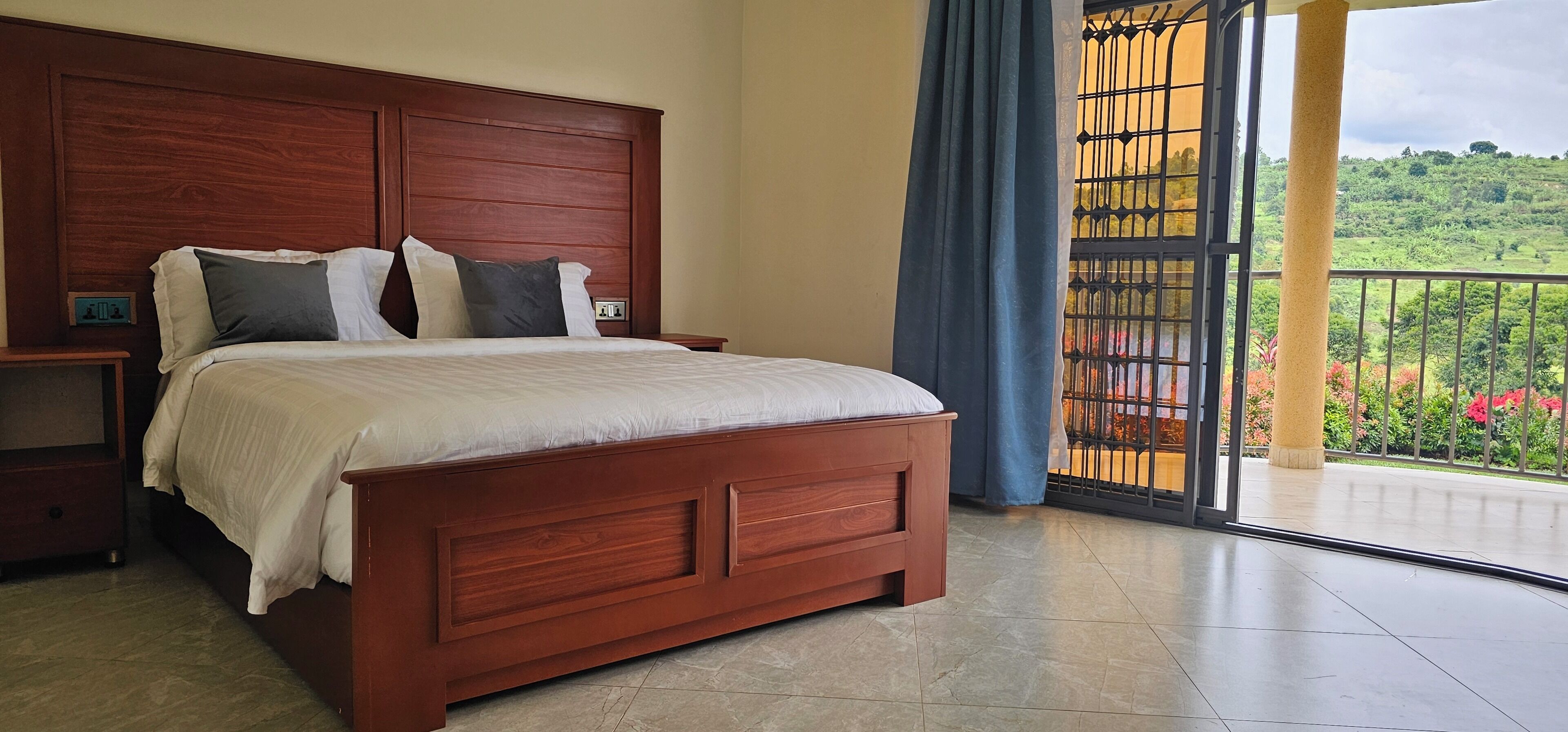 A bedroom with a wooden bed, a nightstand, and a large window with a view of greenery.