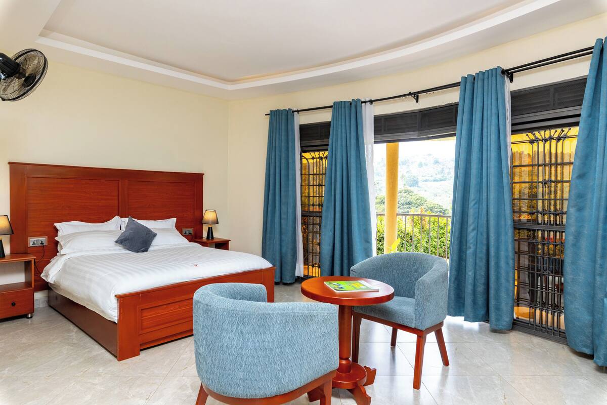 A hotel room with a wooden bed, two blue armchairs, a small table, and a balcony with blue curtains.