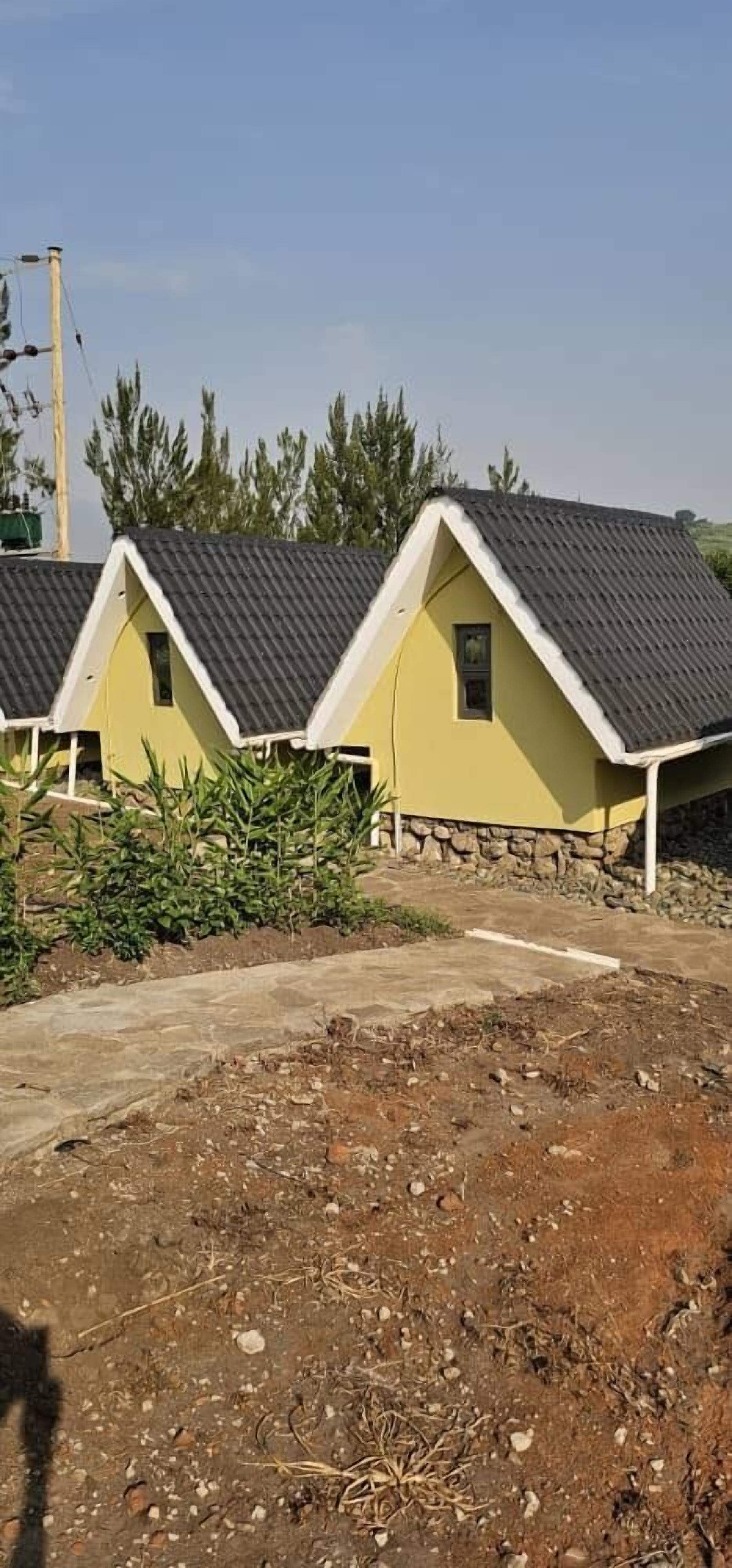 A row of yellow cottages with dark roofs and white trim, surrounded by greenery and a dirt path.