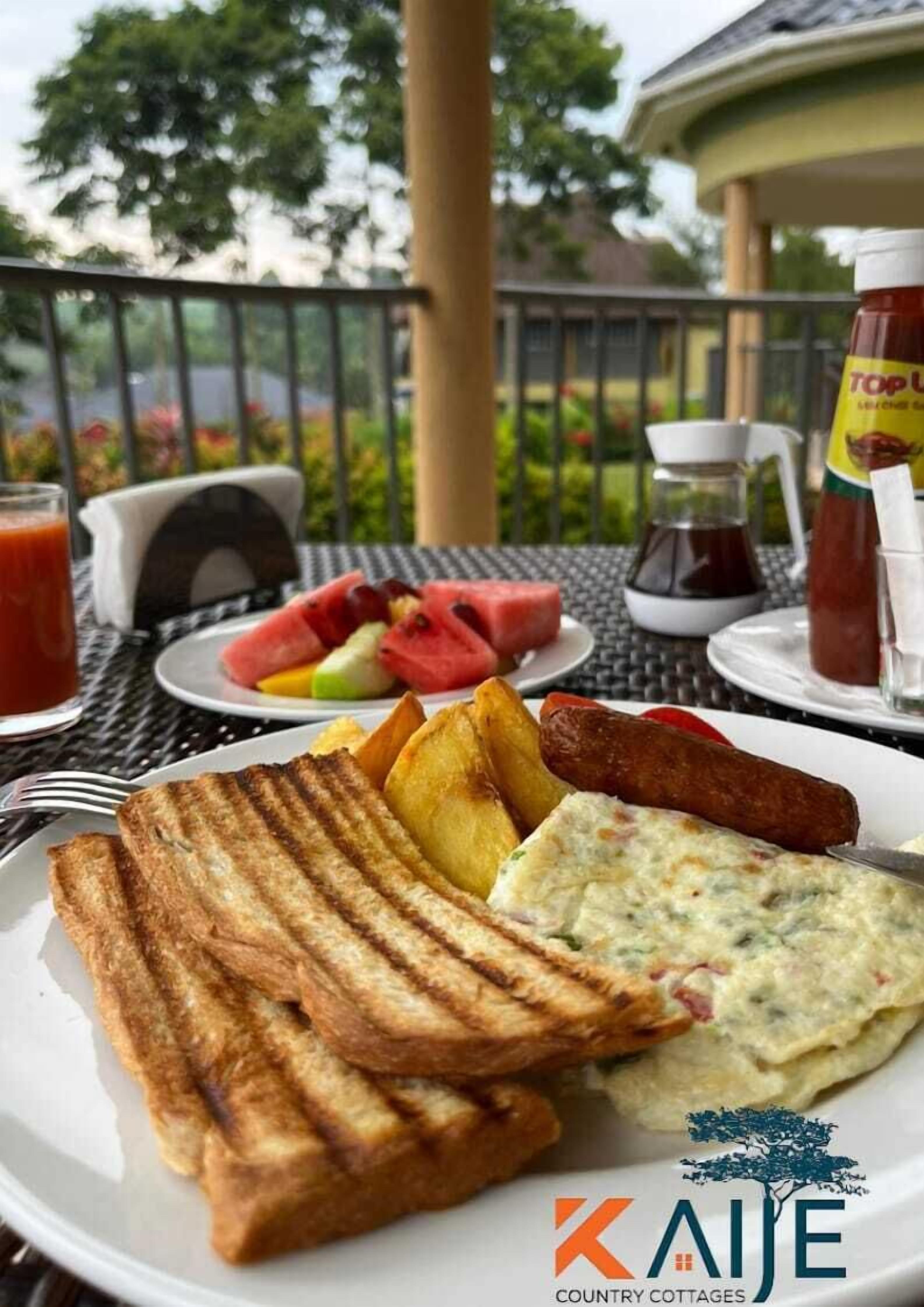 A breakfast plate with toast, eggs, and sausages, accompanied by a glass of juice and a bottle of ketchup, set on an outdoor table with a view of greenery.