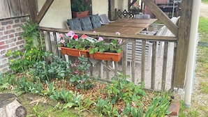 A wooden deck with a bench and hanging flower pots, surrounded by a garden with various plants and flowers. - Grand Gîte (Savigny-sur-Braye)