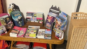 A table with various travel guides and maps, a wicker basket, and a wicker chair. - Grand Gîte (Savigny-sur-Braye)
