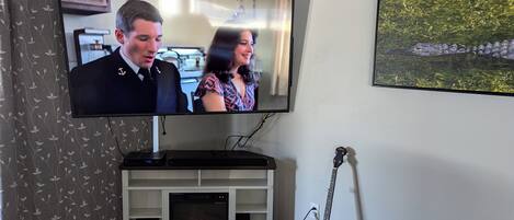Una sala de estar con una televisión de pantalla plana montada en un mueble blanco, una chimenea, una guitarra y un cuadro enmarcado en la pared.