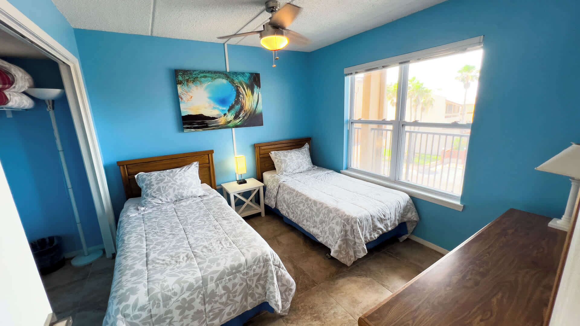 A room with two beds, a ceiling fan, a window with a view, and a painting on the wall.
