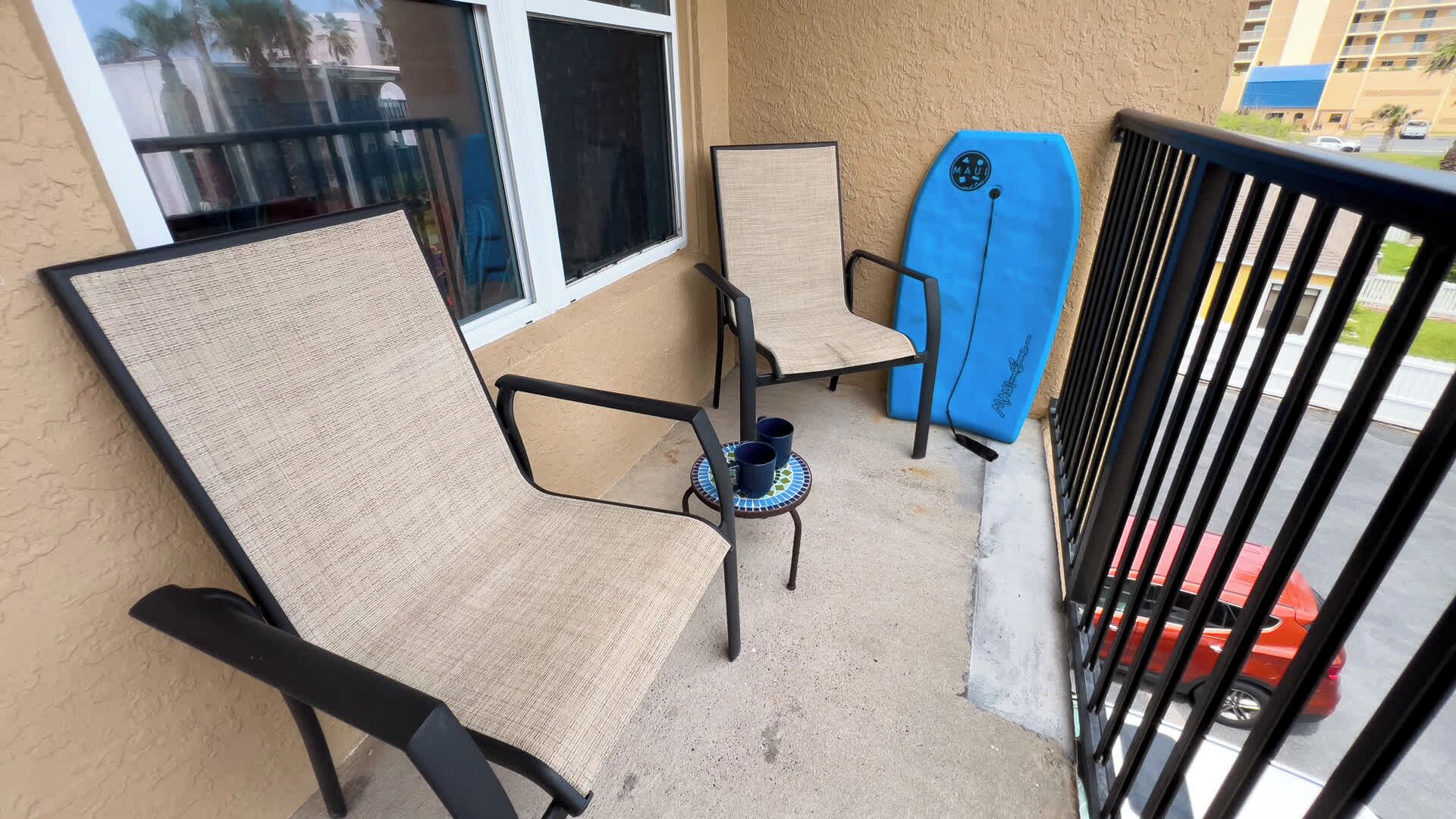 A balcony with two chairs, a surfboard, and a small table with two cups.