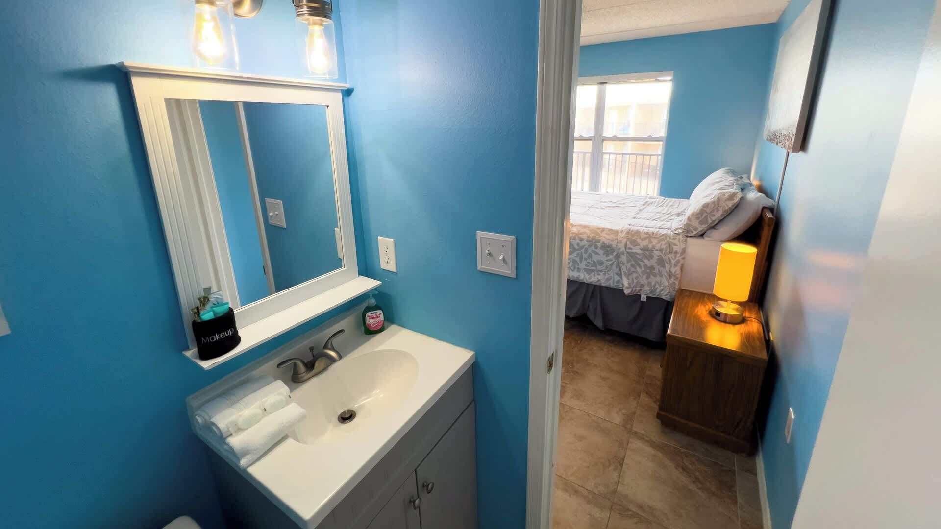 A bathroom with a white sink, a mirror, and a vanity. There is a bed visible through an open door in the adjacent room.