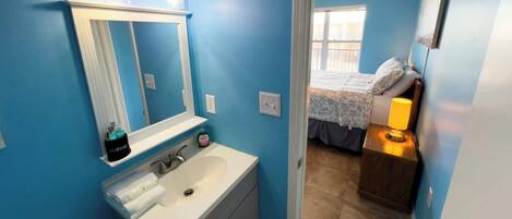 Un baño con lavamanos blanco, un espejo y tocador. Se ve una cama a través de una puerta abierta en la habitación contigua.