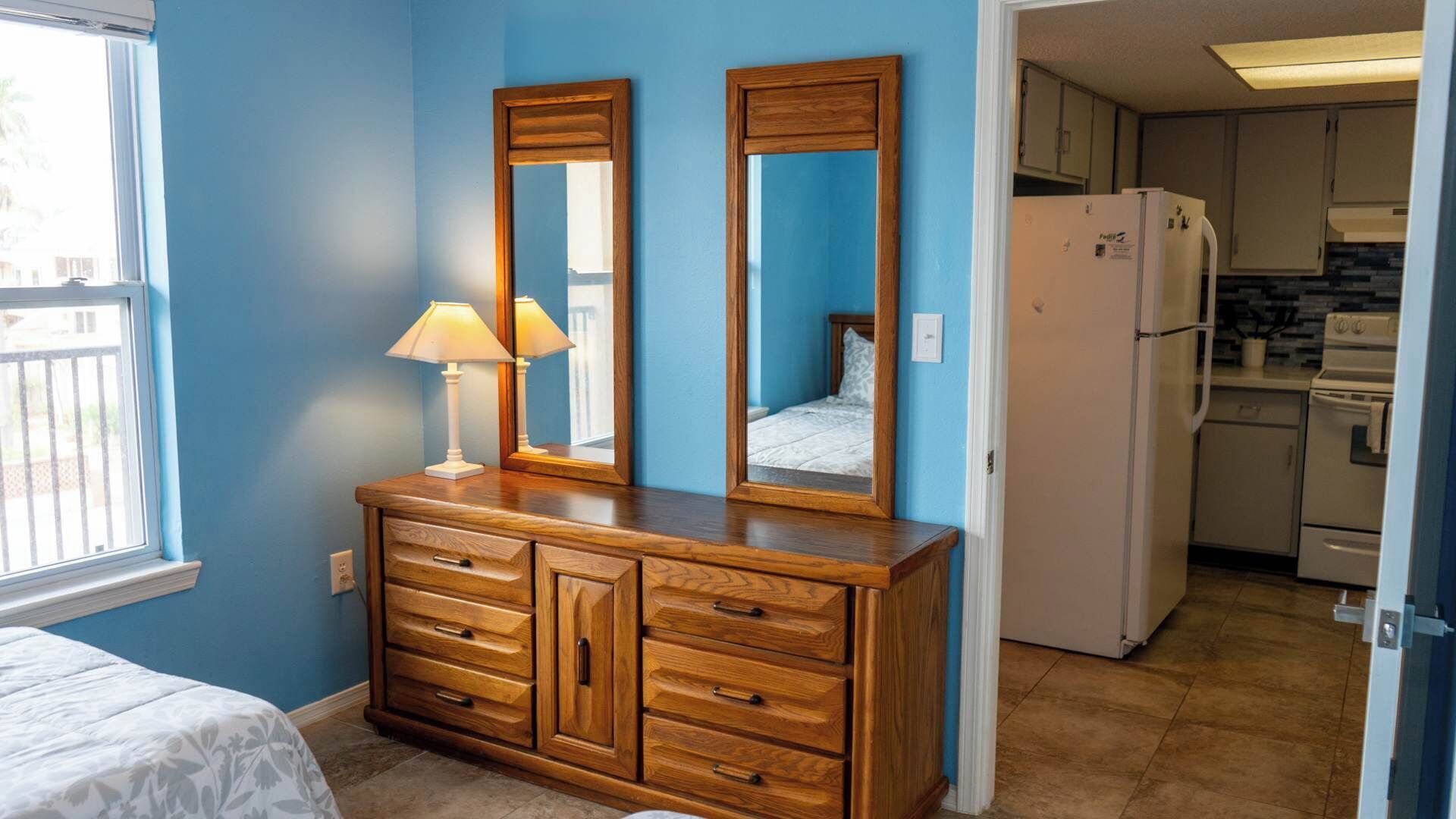 Un dormitorio con un aparador de madera, dos espejos, una lámpara de noche y una ventana.