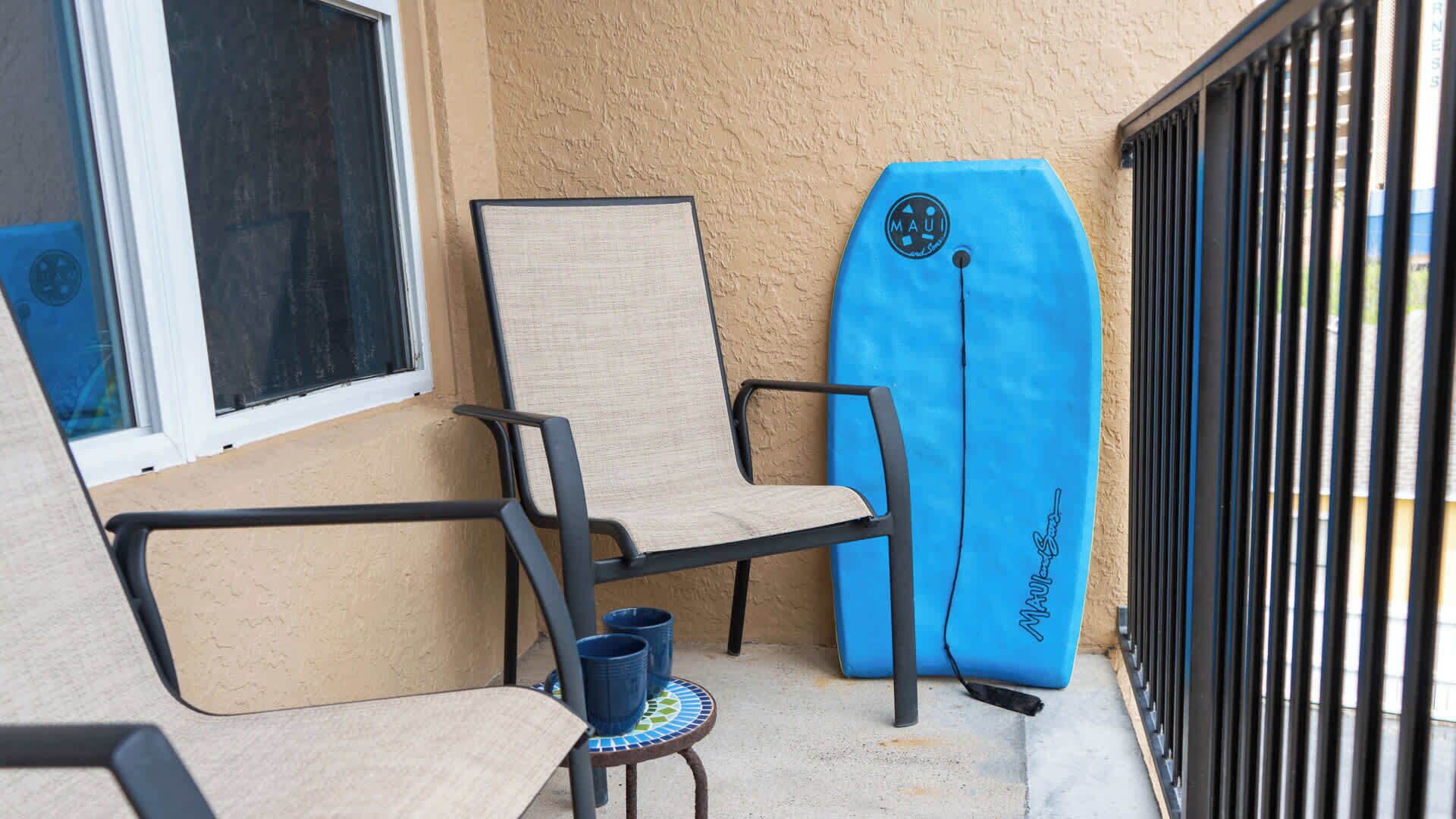 A chair and a surfboard on a balcony with a window and a metal gate.