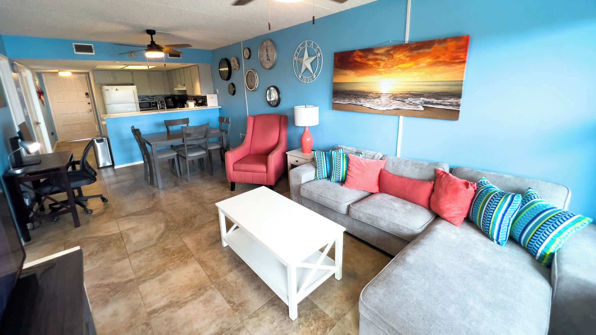 A living room with a gray sofa, red armchair, and a white coffee table. There is a kitchen in the background with a dining table and chairs.