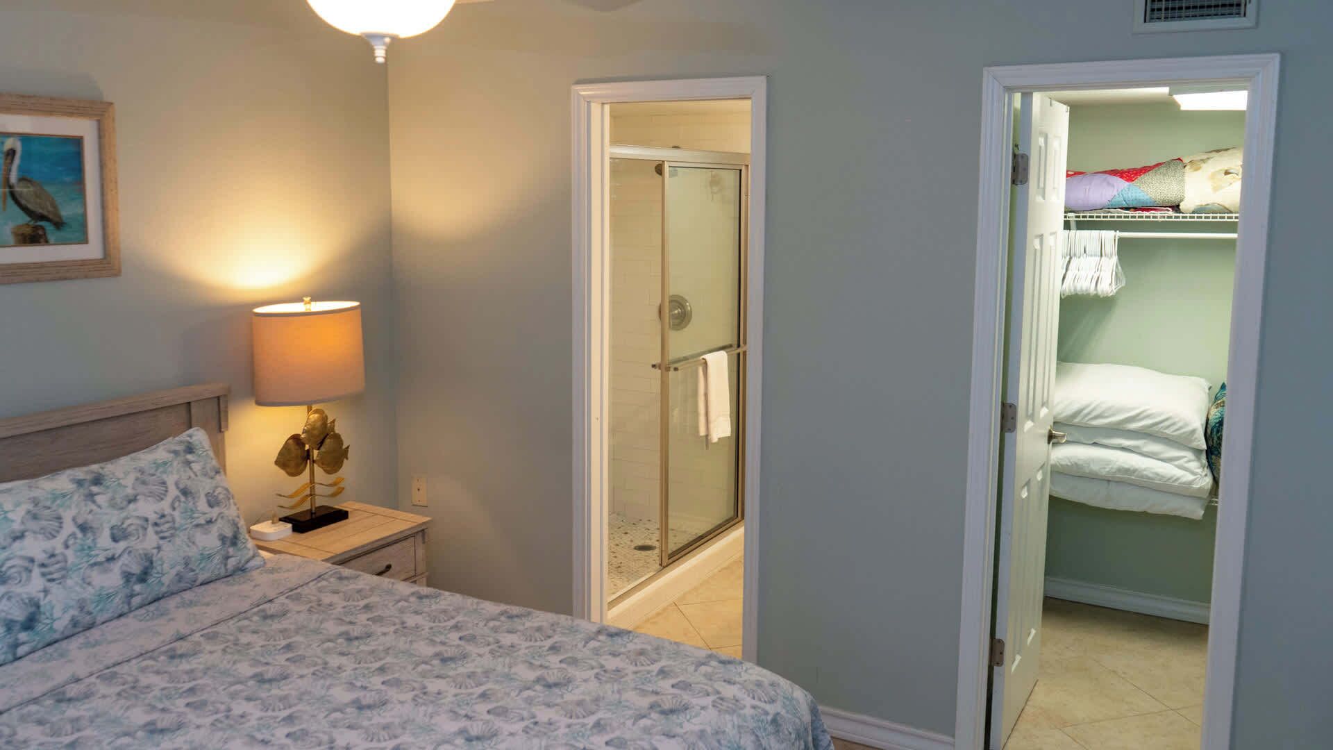 A bedroom with a bed, a nightstand with a lamp, a shower, and a closet with hanging clothes.