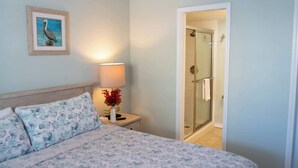 A hotel room with a bed, a nightstand with a lamp and vase, a framed picture on the wall, and a glass-enclosed shower area. - Updated - Comfy Inside - Beach 50 Yards Away (South Padre Island)
