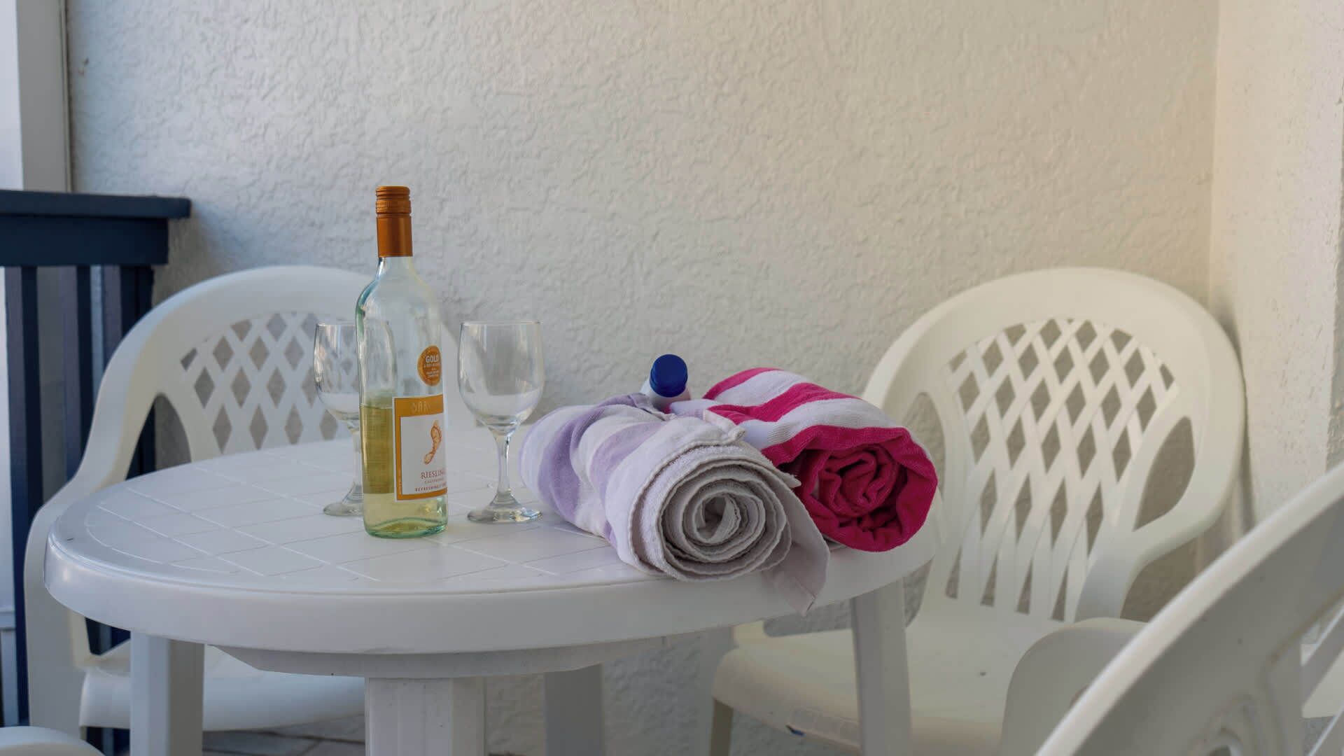 Una mesa blanca con una botella de vino, dos copas y una toalla enrollada con una rosa.