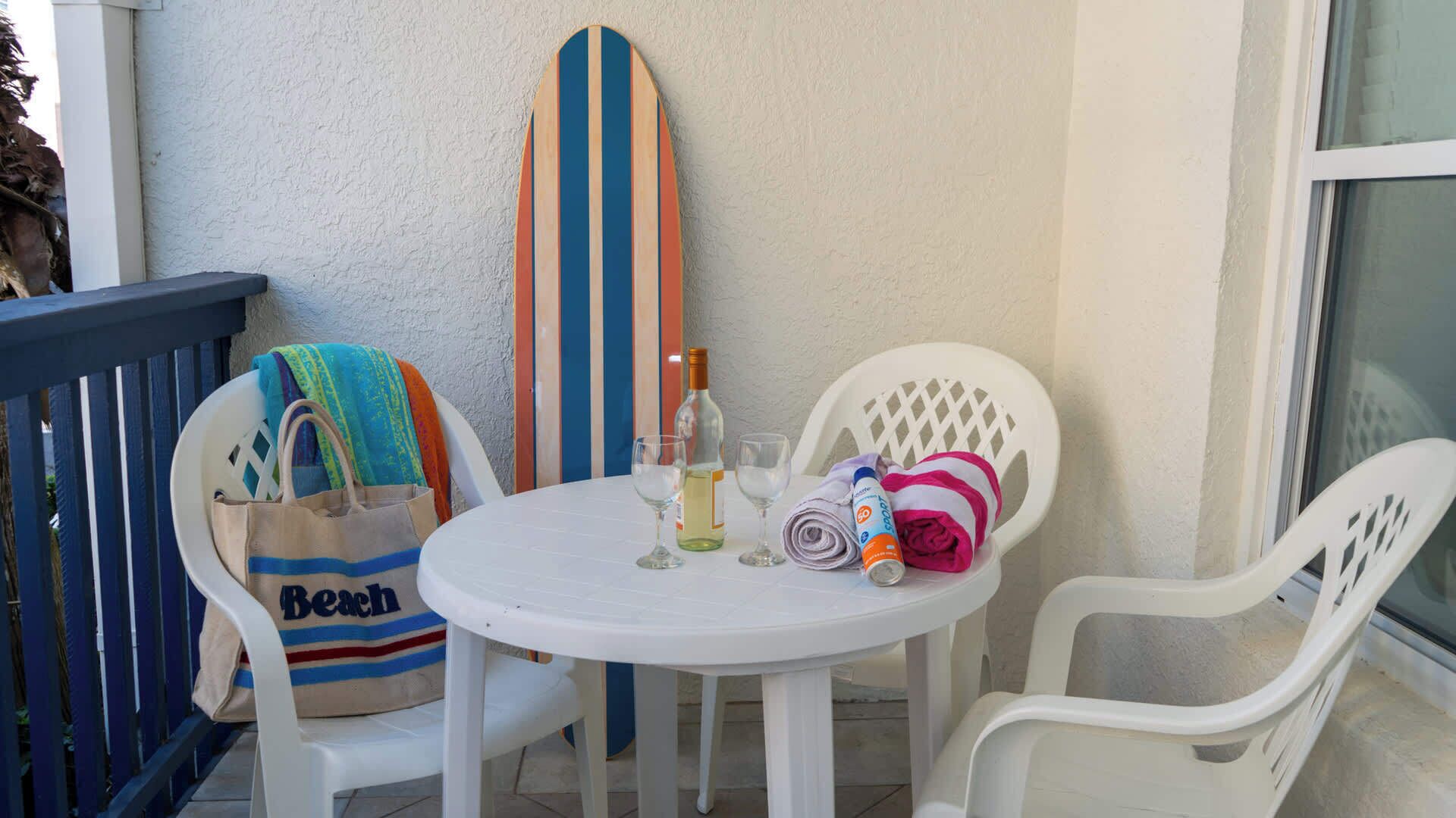 A small outdoor seating area with a round table, two white plastic chairs, a surfboard, a beach towel, a bag, and a bottle.