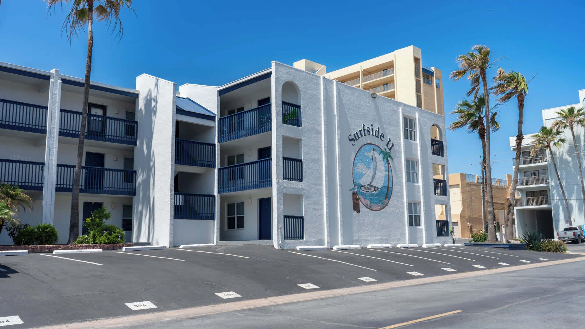 Un hôtel situé en bord de mer, doté de nombreux balcons, d’un parking et de palmiers.