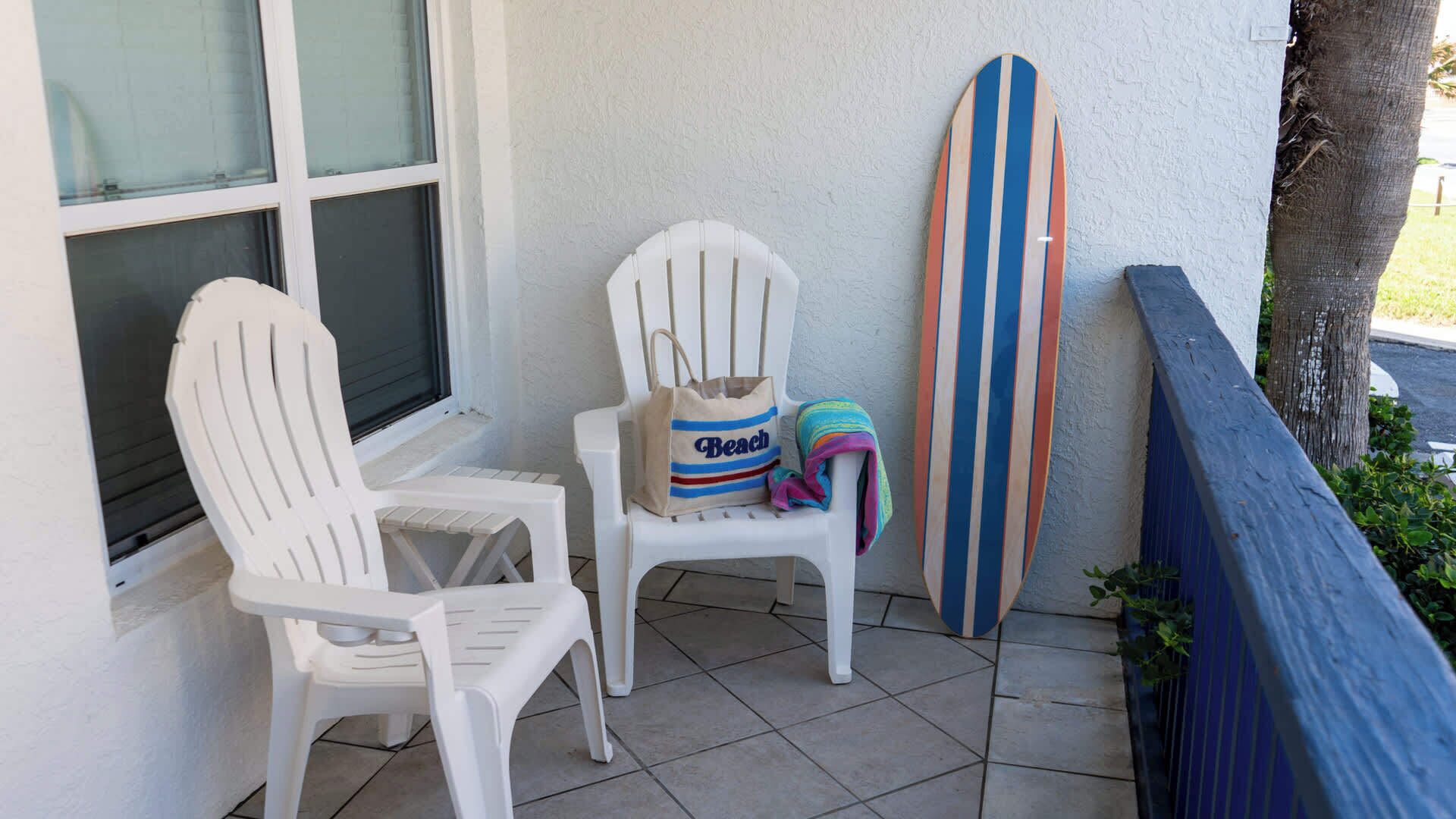 Une terrasse avec deux chaises en plastique blanches, un sac à l’effigie d’une plage et une planche de surf rayée appuyée contre un mur.