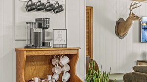 Ein hölzerner Konsolentisch mit einer Kaffeemaschine und Tassen, ein an der Wand befestigtes Schild mit motivierendem Text und ein aufgehängter Hirschkopf.