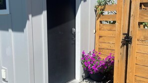 Un bâtiment gris avec une porte noire, un portail en bois et un pot de fleurs contenant des plantes aux fleurs violettes.