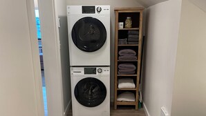 Une buanderie équipée de deux machines à laver à chargement frontal superposées, d’une étagère en bois où sont pliées des serviettes et d’un radiateur fixé au mur.