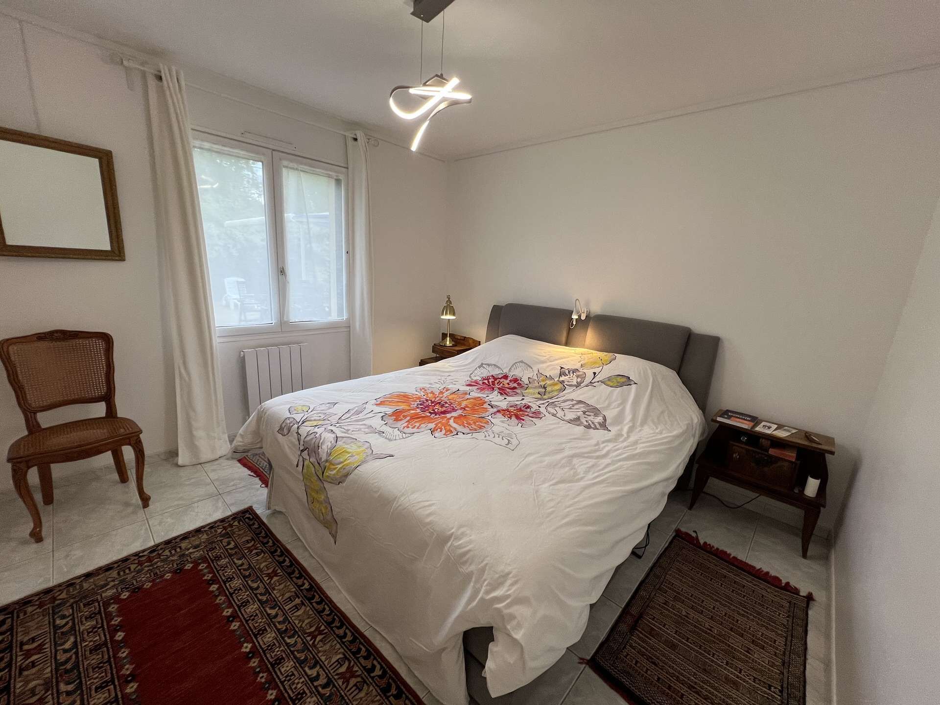 Une chambre à coucher comprenant un lit, une chaise, une table de chevet, une lampe, une fenêtre avec des rideaux et un radiateur.