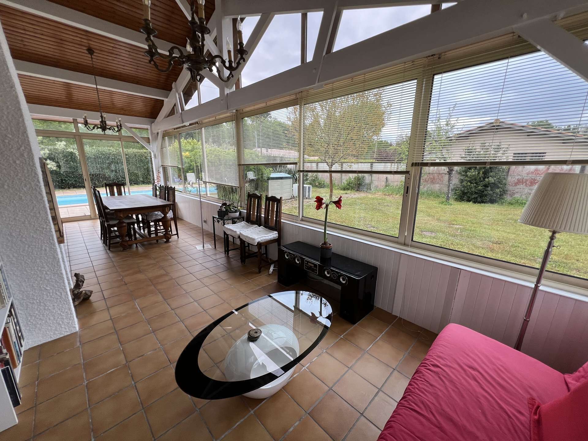 A spacious living area with a dining table, chairs, a glass coffee table, and a red sofa. Large windows provide a view of the outdoors.