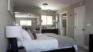 A bedroom with a bed, bedside table, and a nightstand. There is a window with curtains, a door, and a mirror on the wall.