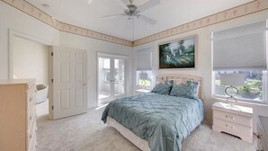 Une chambre à coucher comprenant un lit, une table de chevet, une commode, un ventilateur de plafond et une fenêtre avec des stores.