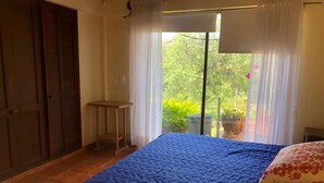 Une chambre à coucher avec un lit, une petite table, une fenêtre avec des rideaux et une vue sur la verdure à l’extérieur.