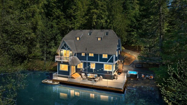 A two-story house with a deck, a hot tub, and a dock extending into a lake, surrounded by dense forest. - Hillside Cottage: Private Pond, Hot Tub, Game Room by Hurricane Ridge, ONP (Port Angeles)