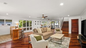 A spacious living room with hardwood flooring, a sofa, a coffee table, and a TV. There is a kitchen area with a bar and stools, and a staircase leading to another level. - 4Br 4.5 Ba Punahele House with Ocean and Mountain Views, Beach Gear, Near Queens Pond (Princeville)