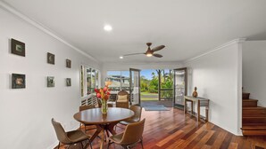 A spacious room with a wooden floor, a round dining table with chairs, a ceiling fan, and a view of a balcony with outdoor furniture. - 4Br 4.5 Ba Punahele House with Ocean and Mountain Views, Beach Gear, Near Queens Pond (Princeville)