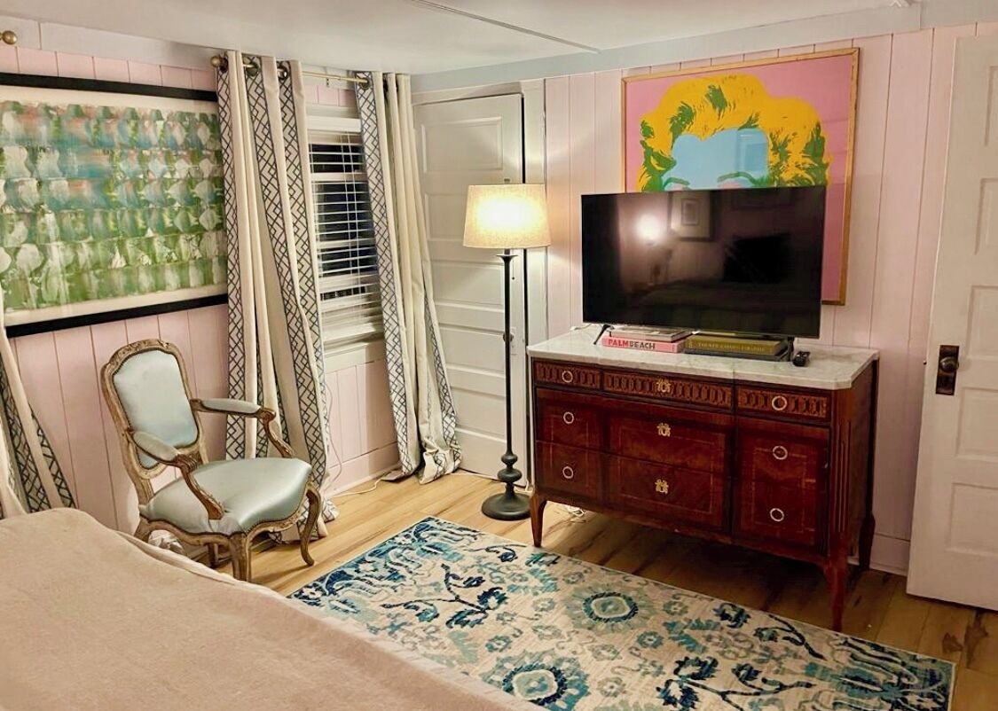 A room with a bed, a chair, a TV on a wooden cabinet, a lamp, and a patterned rug.