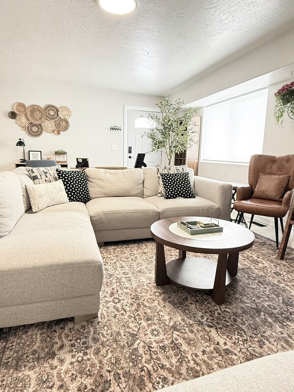 A living room with a beige sectional sofa, a round coffee table, a brown leather armchair, a potted plant, and a wall-mounted woven art piece.