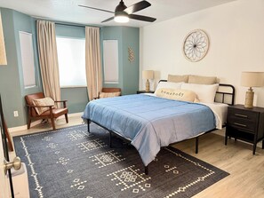 A bedroom with a bed, nightstands, a chair, a ceiling fan, and a decorative wall piece.
