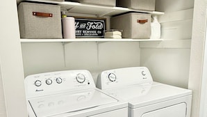 A laundry room with two white washers and dryers, storage boxes on shelves, and a sign for a dry cleaning service. - Restful Nest A 3bd&2ba in Midtown Anchorage (Anchorage)