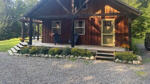 Una cabaña de madera con un porche, dos ventanas, una puerta y una pequeña zona de estar exterior con sillas.