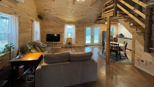 Interior de una cabaña de madera con sofá, televisión, comedor y una escalera.