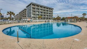 Une grande piscine avec un escalier en métal, entourée d’une zone sablonneuse et d’un immeuble à plusieurs étages doté de balcons.