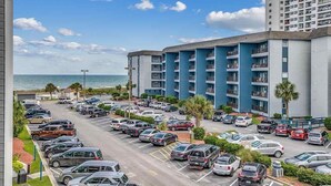 Un hôtel à plusieurs étages avec un parking à l’avant, une vue sur la plage et un grand immeuble en arrière-plan.