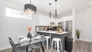 A modern kitchen with white cabinets, a stainless steel refrigerator, and a glass dining table with grey chairs. - Spruce Ave - Parking - Sleeps 4

 (Edmonton)
