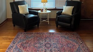 Una sala de estar con dos sillones de cuero negro, una mesita redonda con lámpara, un tapete estampado y un ventilador de techo.