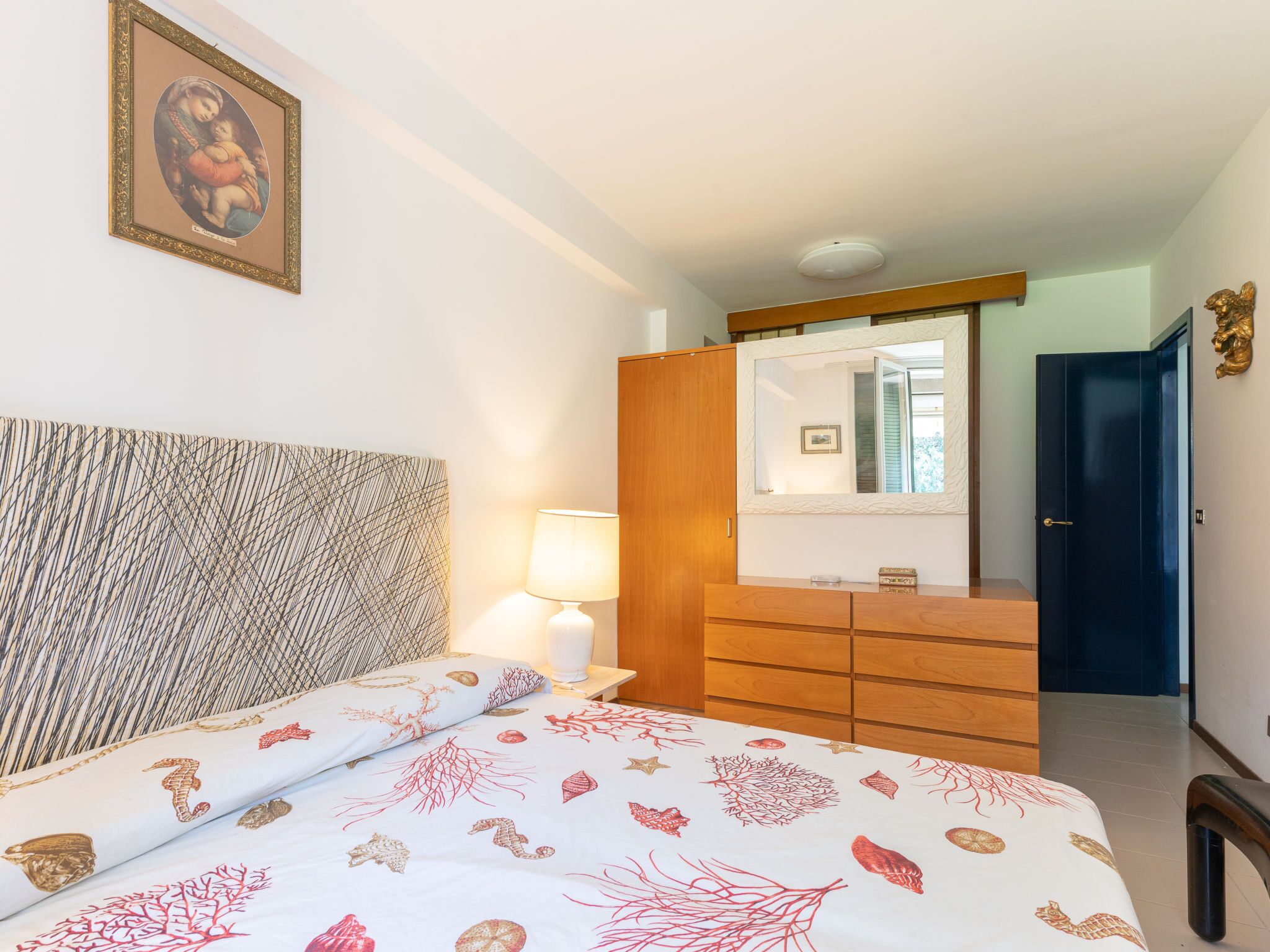Een slaapkamer met een bed, nachtkastje, dressoir, spiegel en een deur.