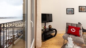 Un balcon avec vue sur la mer, un meuble TV noir, un fauteuil gris avec un coussin rouge et une théière blanche posée sur un plateau.