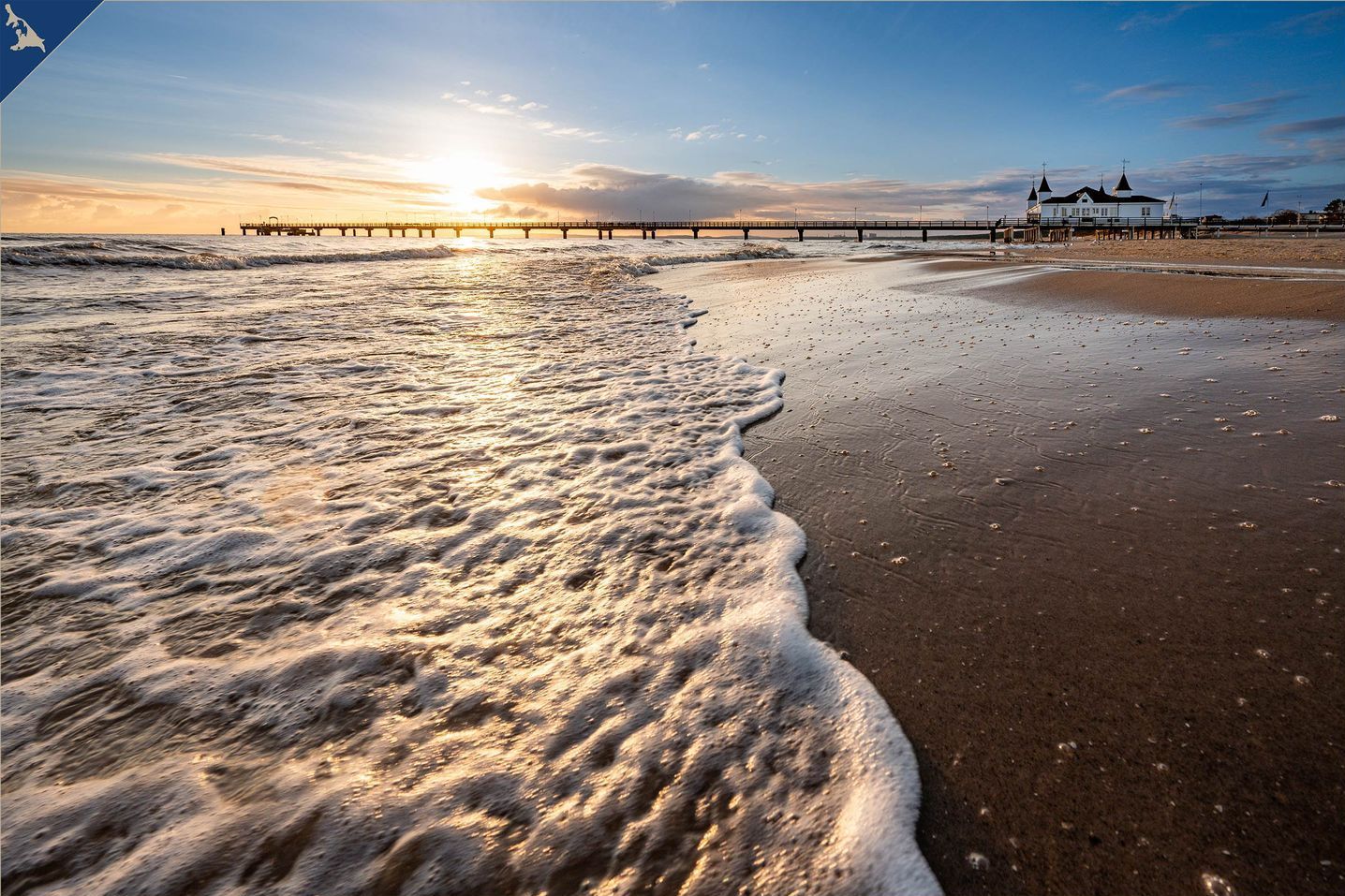 A beach with waves, a pier, and a building with a white facade and multiple spires.