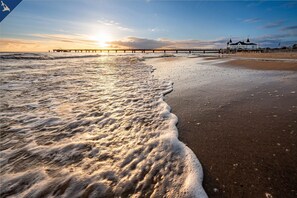Ein Strand mit Wellen, einem Steg und einem Gebäude mit weißer Fassade und mehreren Türmen.