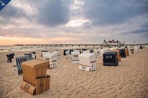 Una spiaggia con file di sedie a rotoli e lettini prendisole, un molo che si protende verso il mare e un edificio con tetti verdi sullo sfondo.