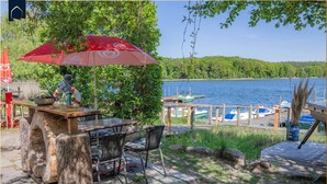A wooden table with chairs, a red umbrella, and a view of a lake with boats.